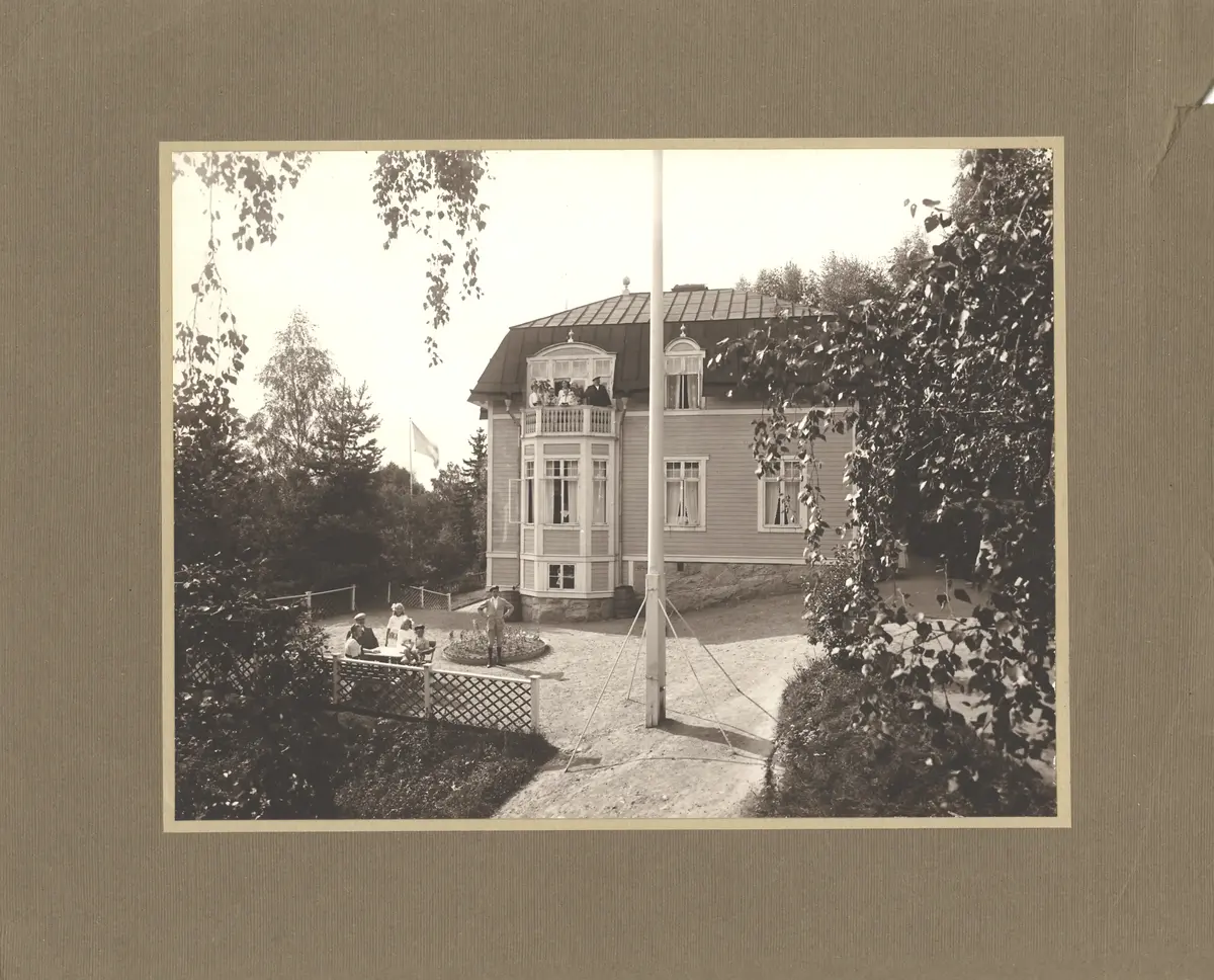 Villa eller sommarnöje; okänd plats. I förgrunden sitter människor runt ett bord; ytterligare en grupp står på villans balkong.