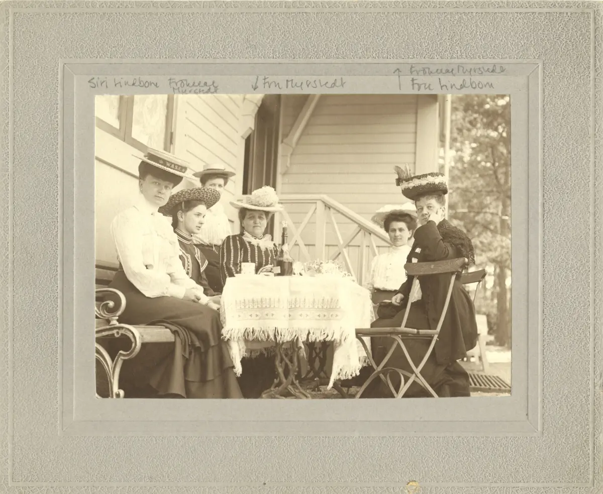 Hyresgäster på Edlunda. 
6 kvinnor sitter runt ett bord på en  veranda. På bordet står kaffekoppar och en flaska. 

Avbildade personer:Siri Lindbom; Fru Myrstedt; Fröken Myrstedt; Fru Lindbom
