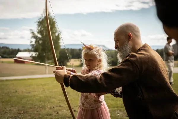 Utendørs sitter en mann ikledd jernalderklær på huk med pil og bue i hånda og viser et barn hvordan pilen skal sitte på buen