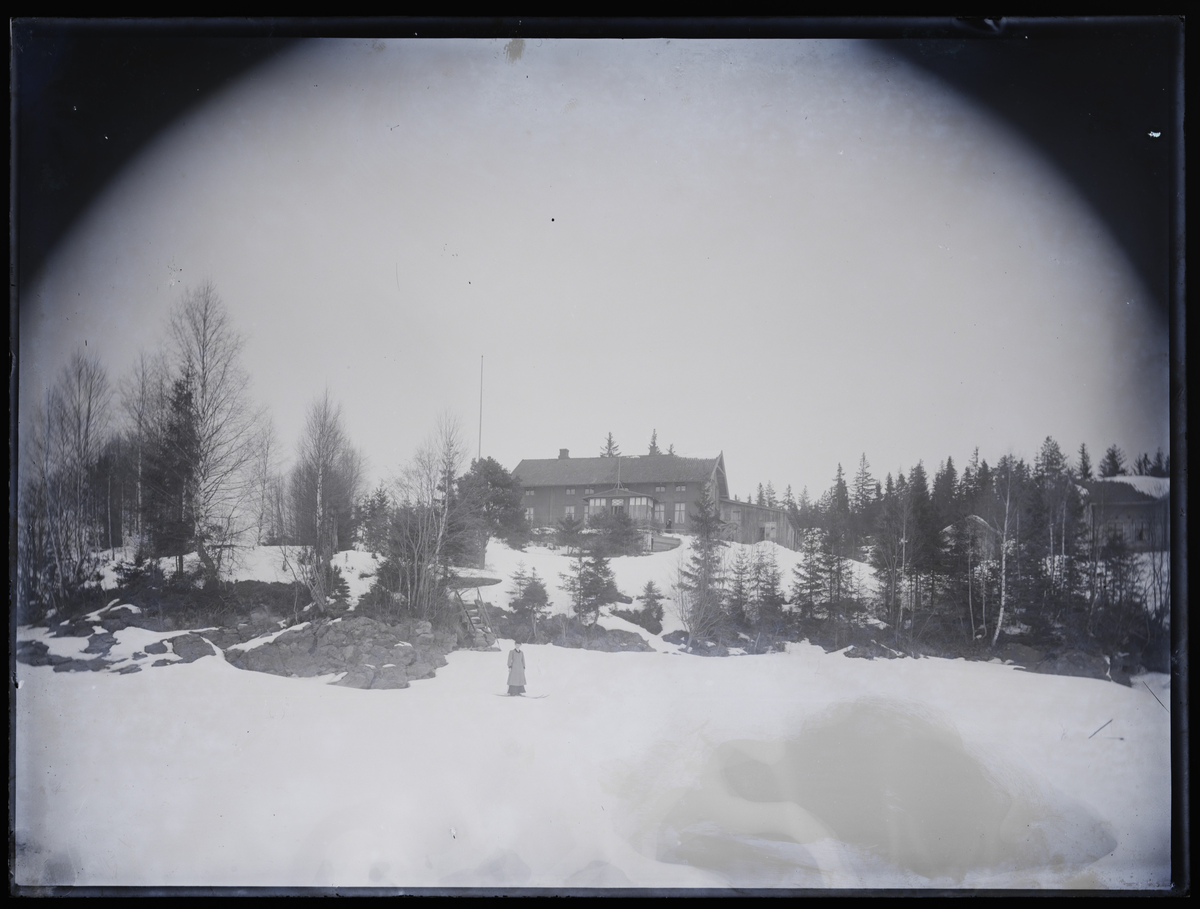 Kvinne på ski i vinterlandskap foran bygning. Antakelig fotografert i Eidsvoll rundt 1908.