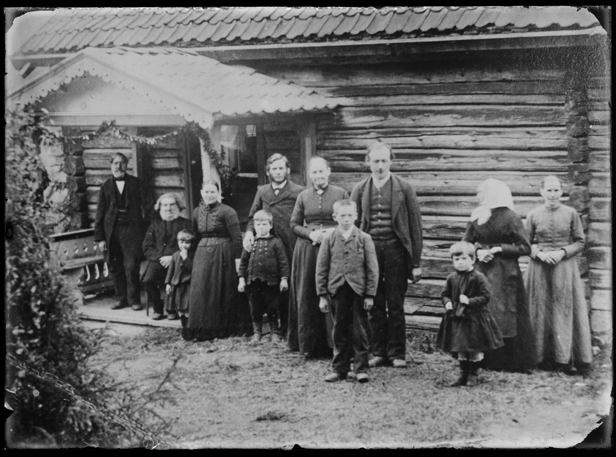 Gruppeportrett med flere generasjoner stående foran et bolighus i laftet tømmer med dekorert inngangsparti.