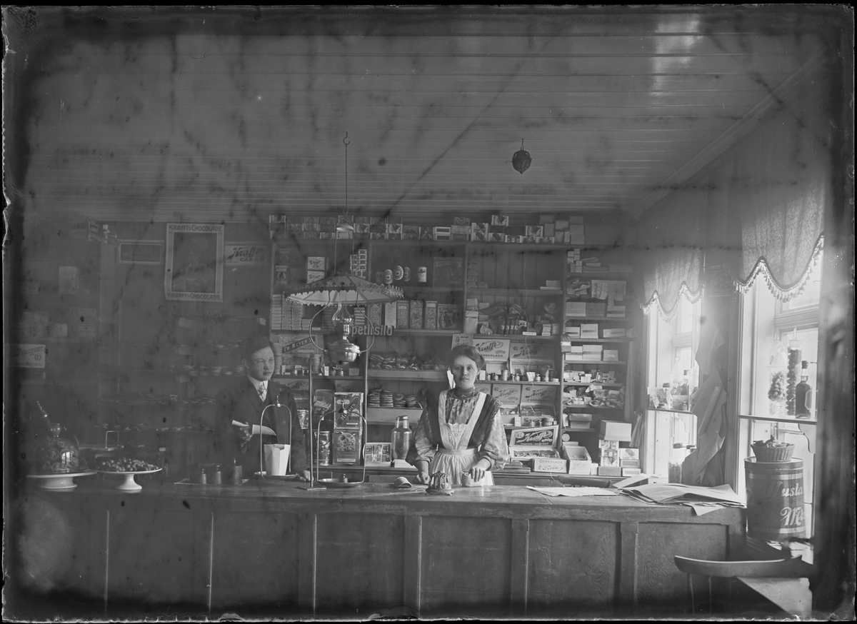 Kvinne og mann står bak disk i en landhandel. Antakelig fotografert i Eidsvoll rundt 1908.