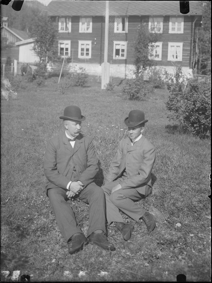 To menn sitter i hage foran et hus. Tekst på baksiden: Paul og Paal. Antakelig fotografert i Eidsvoll rundt 1908.