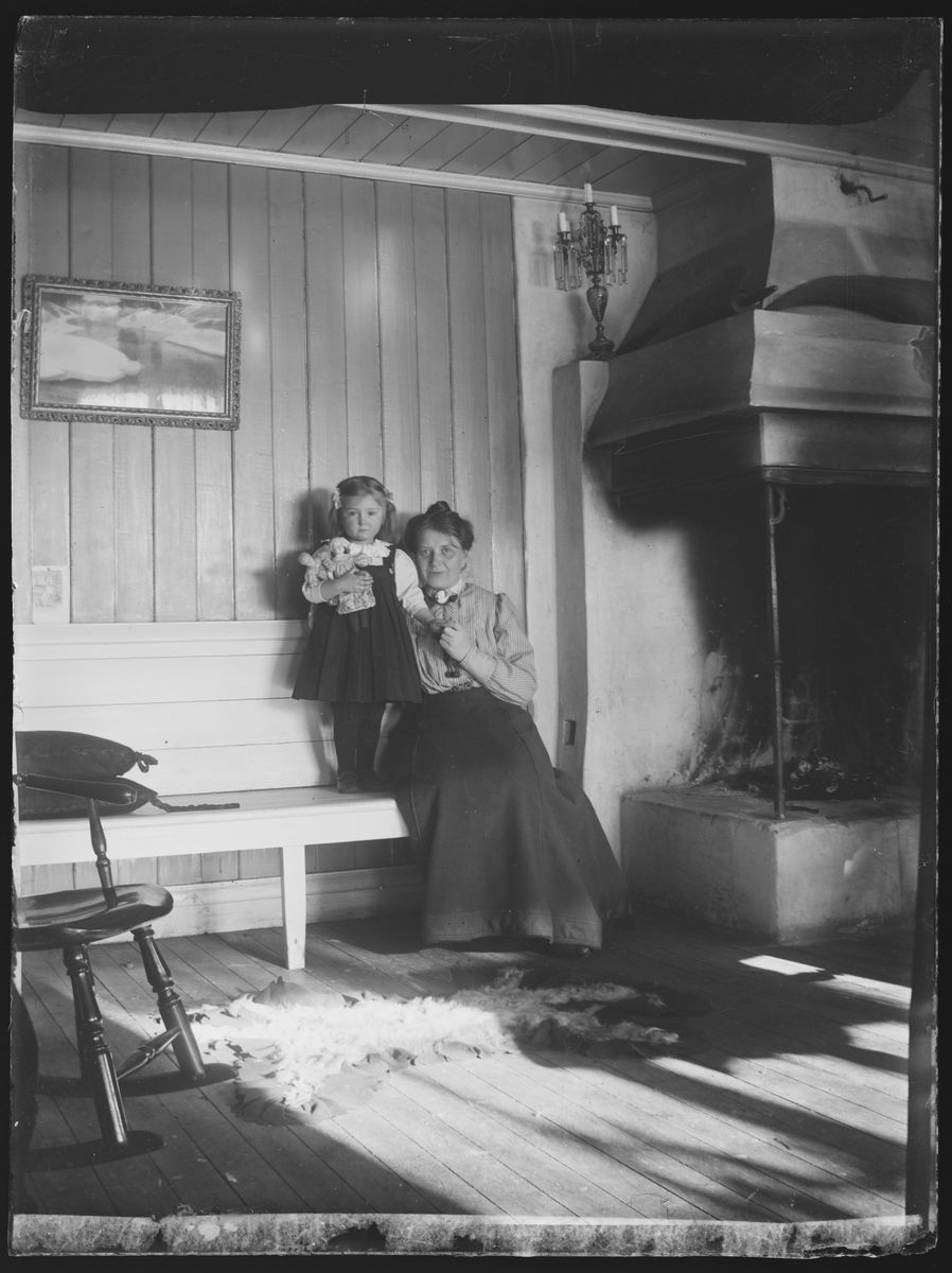 Portrett av mor og datter ved peisen. Tekst på baksiden: Fru Grude Coth med sin datter Aase, 1909.