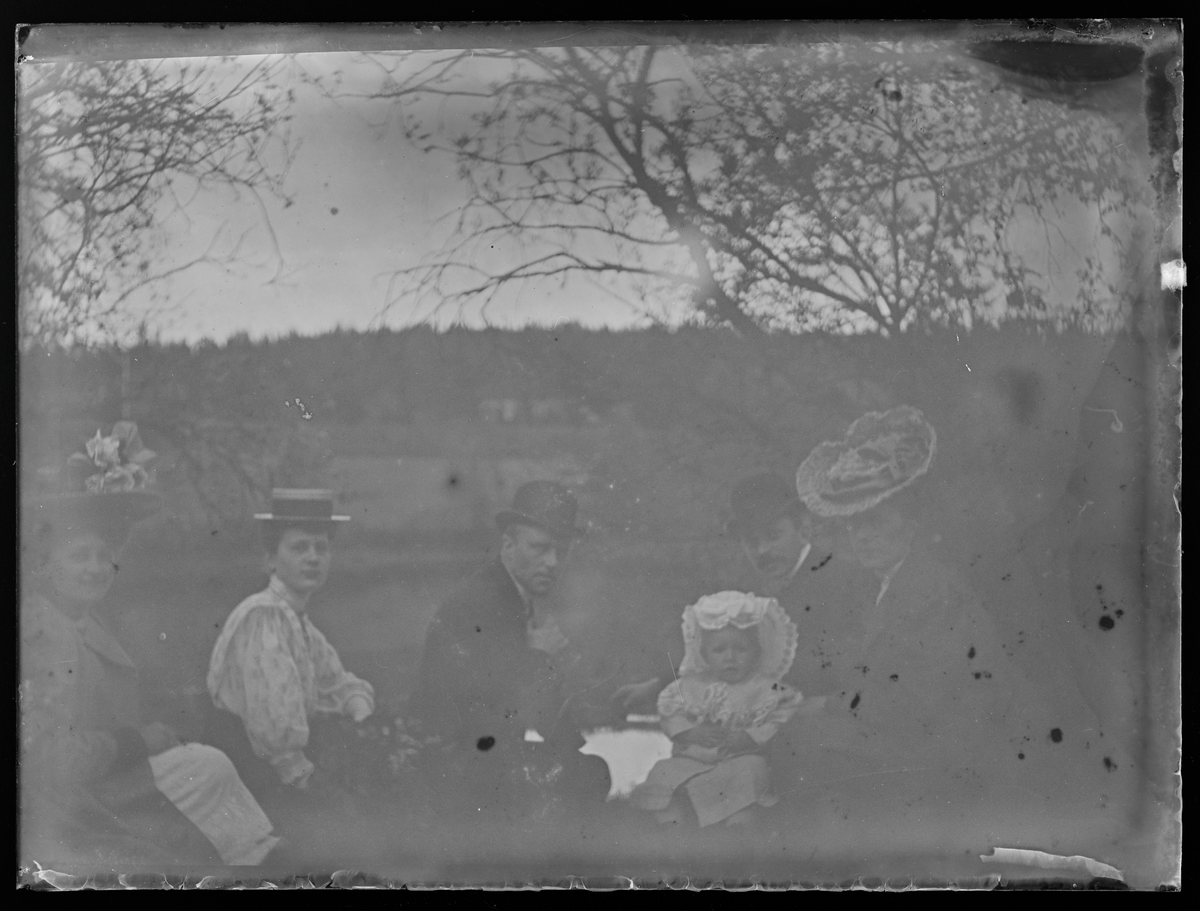 Portrett av kvinner og menn som sitter med en baby i gresset. 

Tekst på baksiden: Lidet møde paa Samfunstangen, 1909.