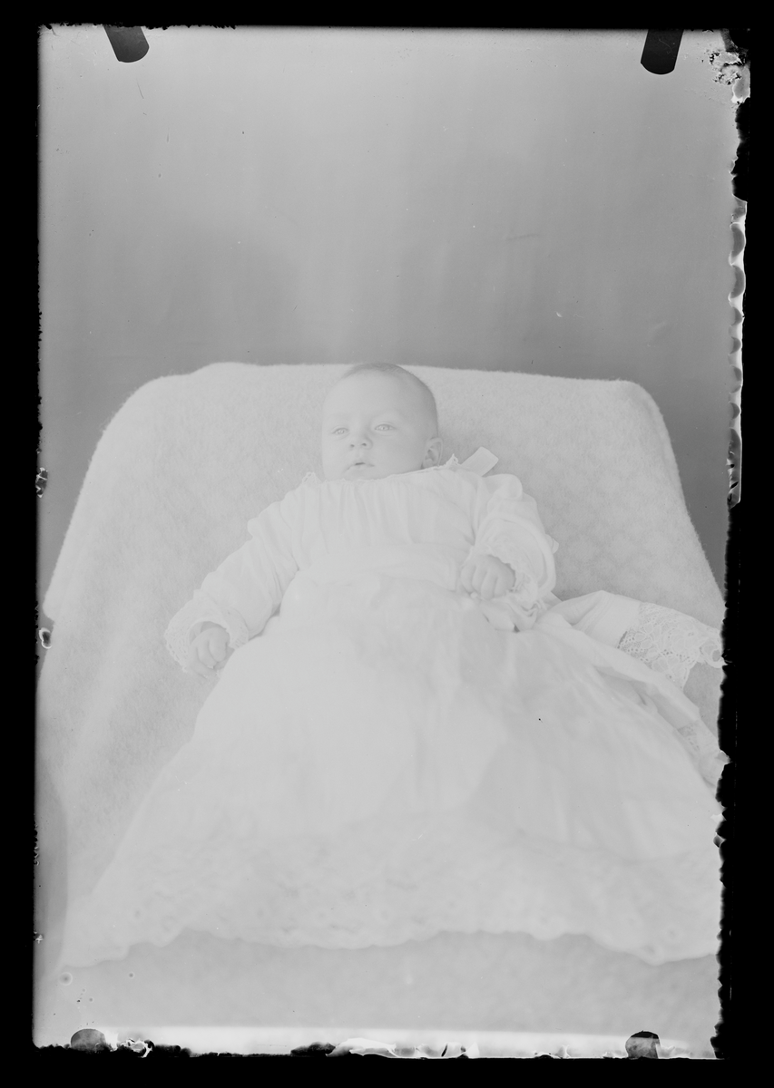 Studioportrett av et spedbarn pyntet i hvit kjole. Antatt barnedåp. Antatt fotografert i Eidsvoll Folkehøgskole omtrent 1908-09.