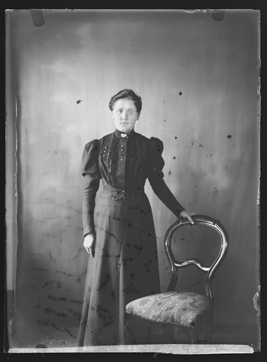 Studioportrett av en kvinne stående ved siden av en stol. Antatt fotografert i Eidsvoll Folkehøgskole 1908-09.