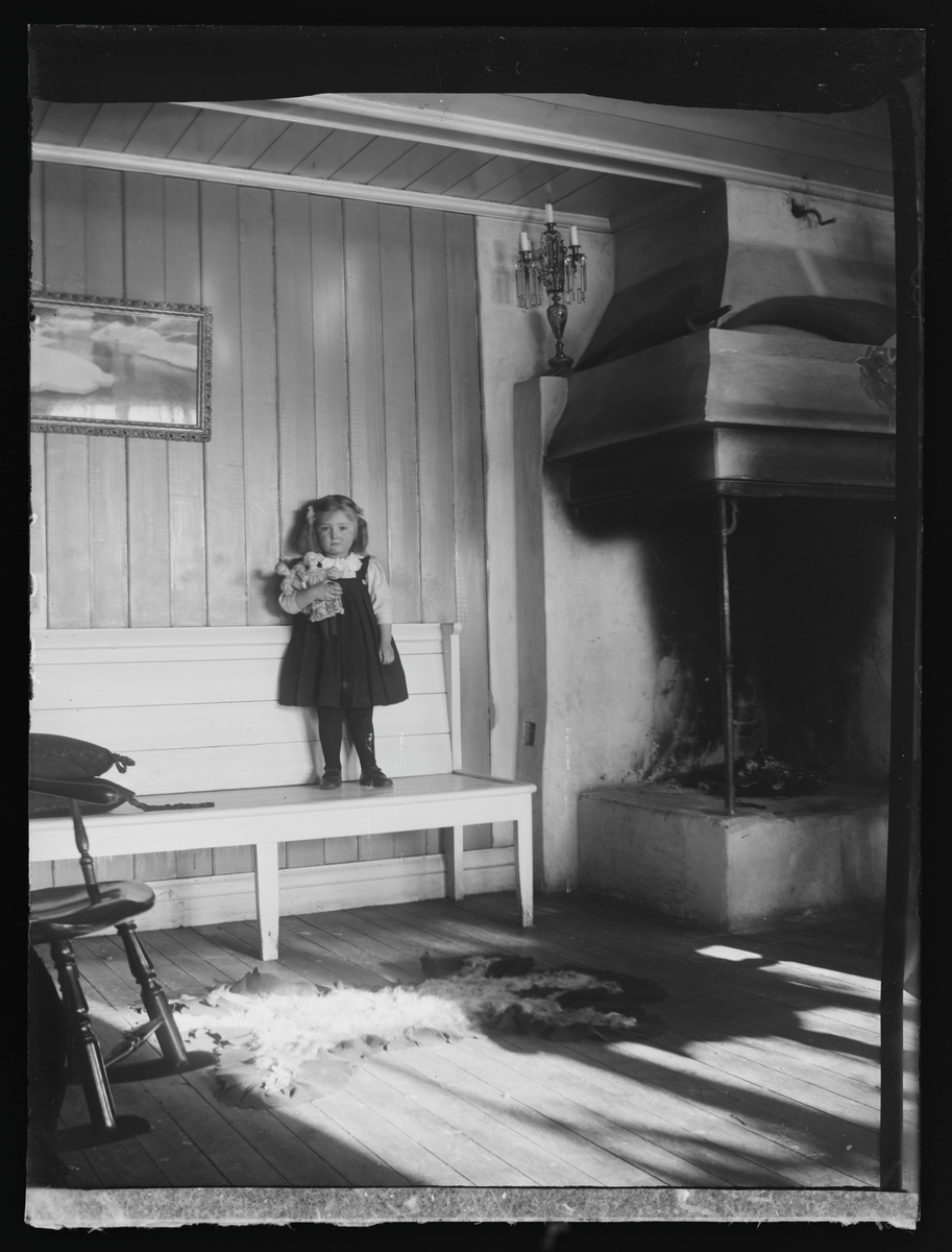 Portrett av jente stående på en benk i en peisstue i Eidsvoll Folkehøgskole fotografert i 1909.

På konvolutt Aase Coth, 471. No. 16. 1909.
