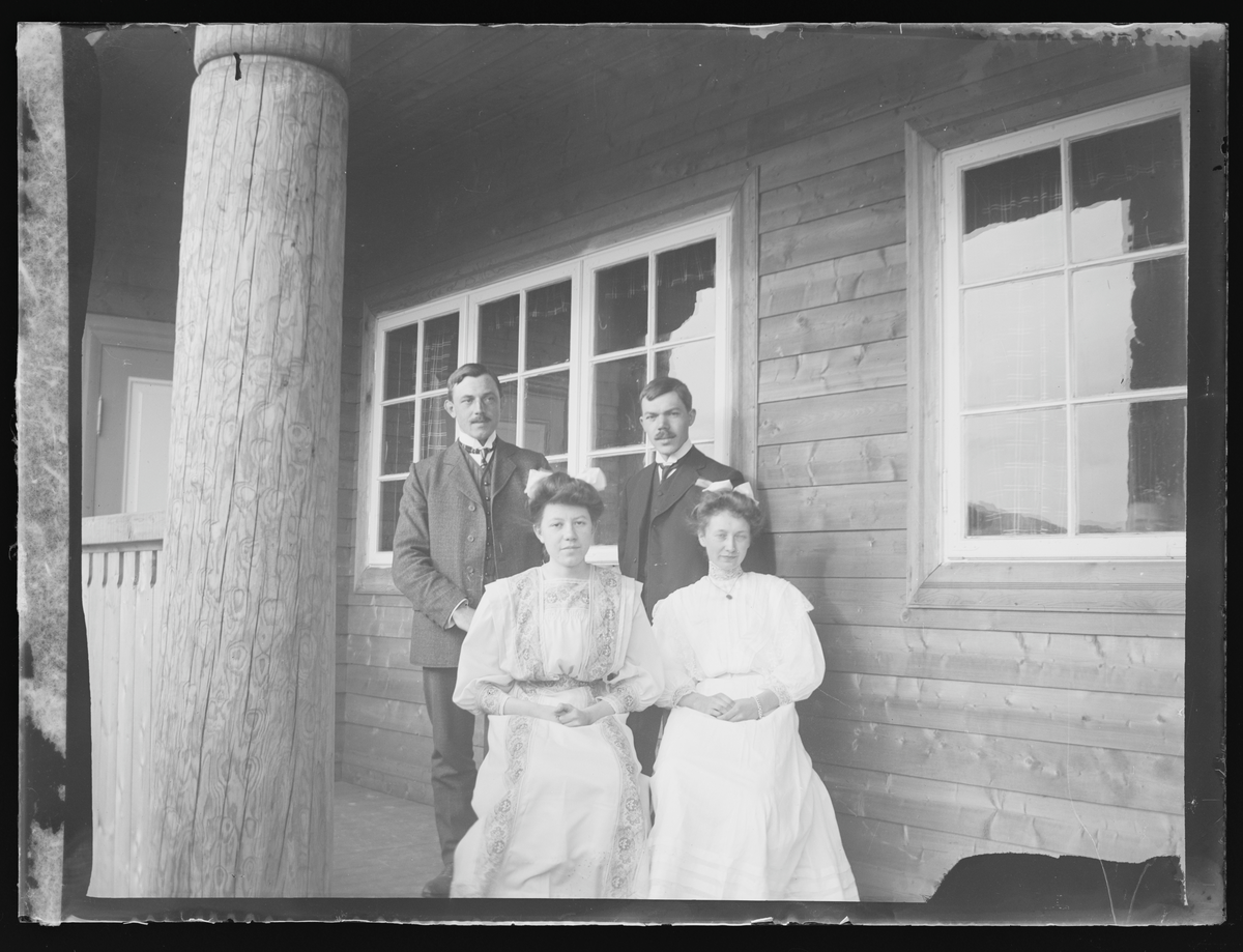 Portrett av to menn og to kvinner på en terrasse.

På konvolutt "Ude paa Verandaen mod vest...K. v. .Olafsby, Borud og Herr Olafsby.,, 472. No. 3. 1909."