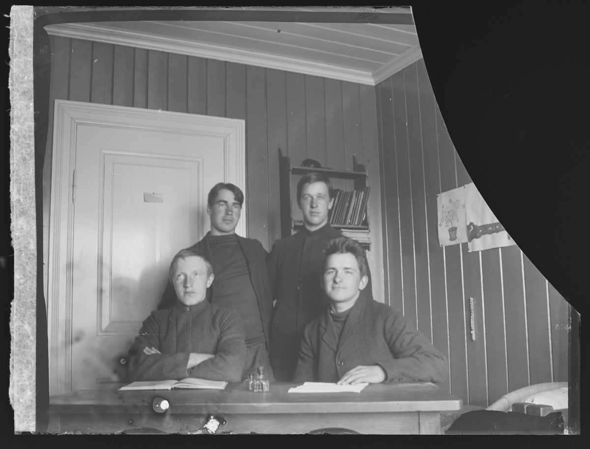 Portrett av fire menn bak en skrivepult i ett av folkehøgskolens soveværelser. Antatt elever ved Eidsvoll Folkehøgskole fotografert i 1909.
Antatt påskrift konvolutt "No 37, 1909, Værels til Sørli og Torsstensen,  Goplen og Nygaard, 476.