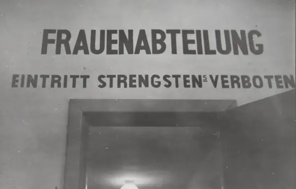 Fotografi svart/hvitt tatt inne i Grini fangeleir, inne i murbygningen ved inngangen til Kvinneavdelingen. Man ser en dør som er åpen og over døra står det "FRAUENABTEILUNG. EINTRITT STRENGSTENS VERBOTEN".
