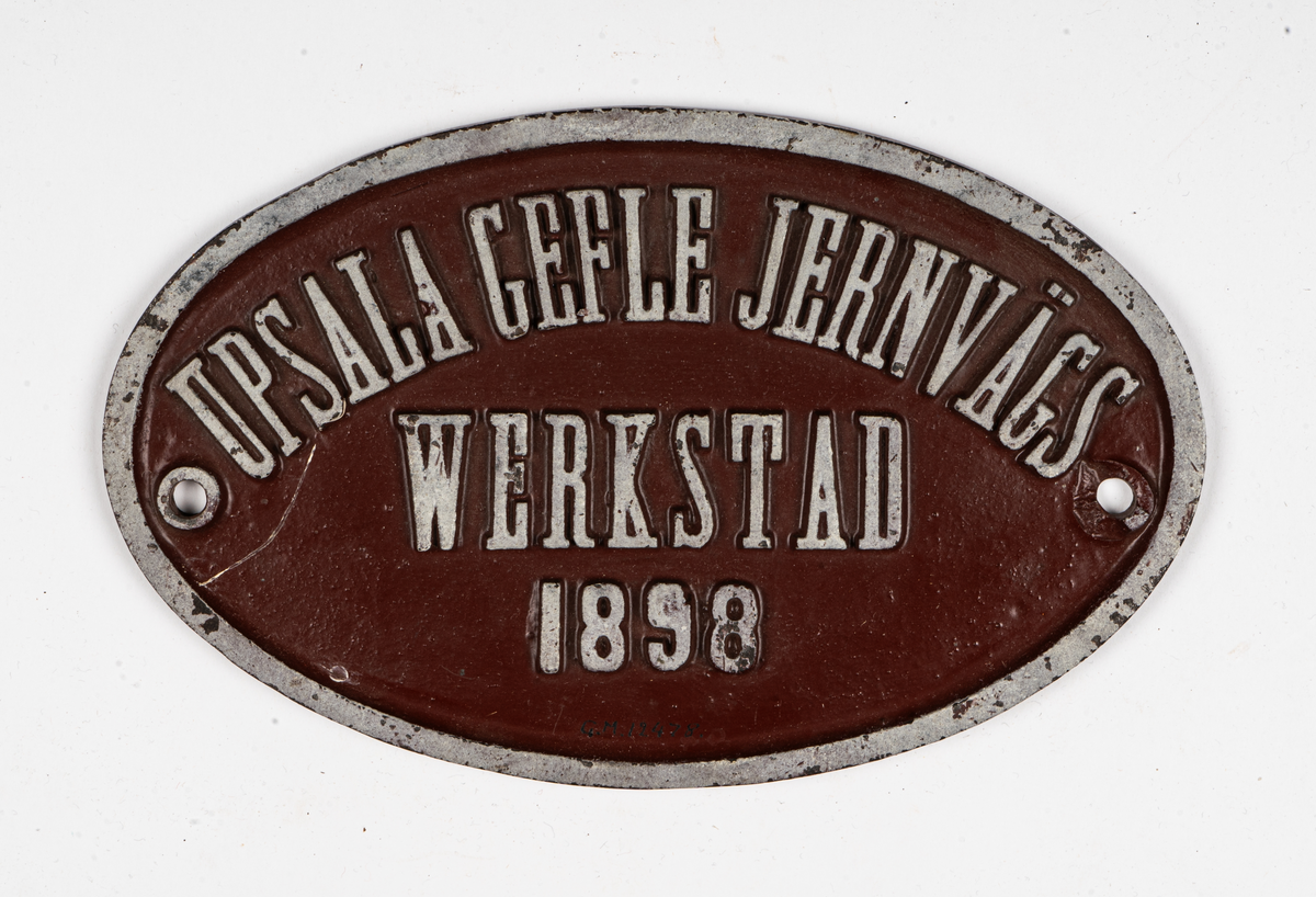 Skylt, fabrikationskylt, oval, av järn, rödmålad, med reliefbokstäver "Upsala Gefle Jernvägs Werkstad 1898". För personvagn.