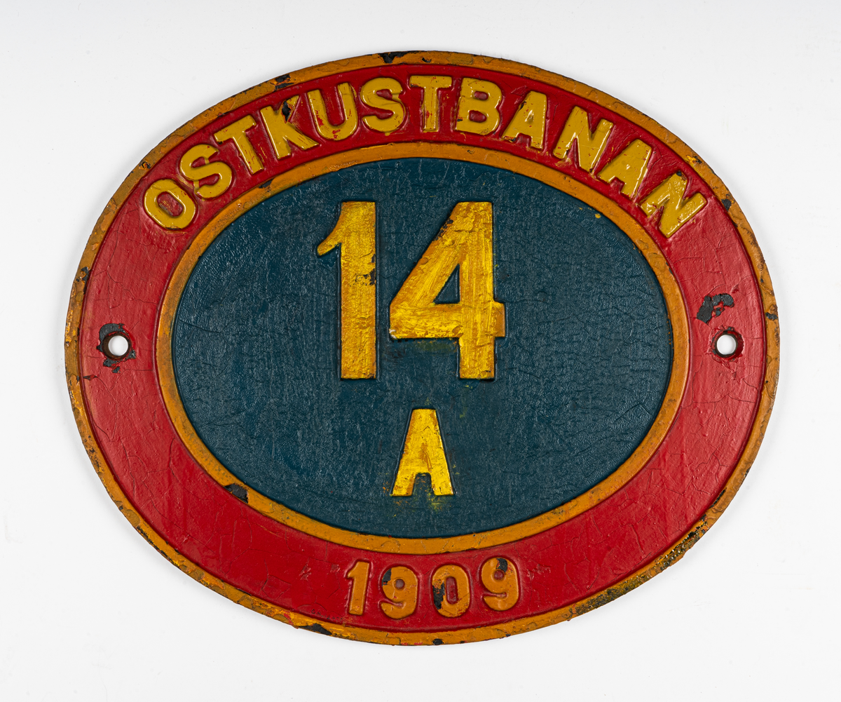 Skylt, lokskylt, för A-lok. Oval av järn, målad rött, blått, guld. Reliefer "Ostkustbanan 14 A. 1909".