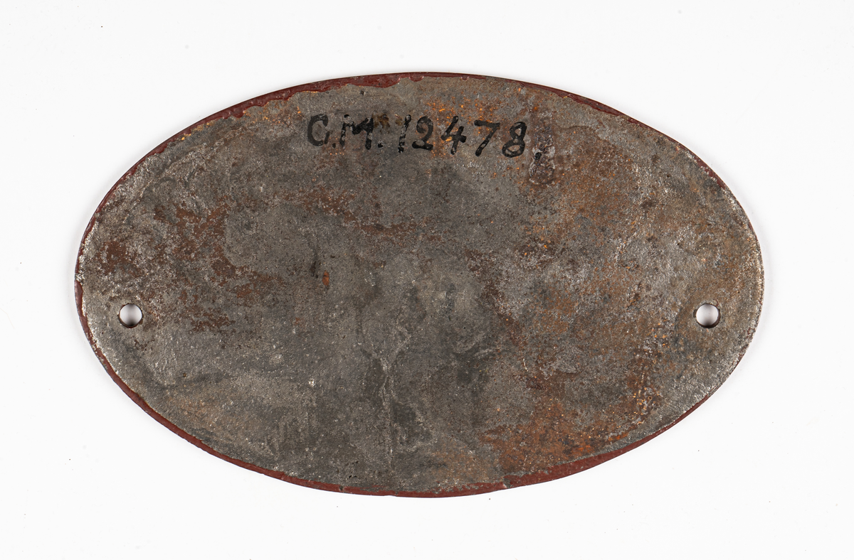 Skylt, fabrikationskylt, oval, av järn, rödmålad, med reliefbokstäver "Upsala Gefle Jernvägs Werkstad 1898". För personvagn.
