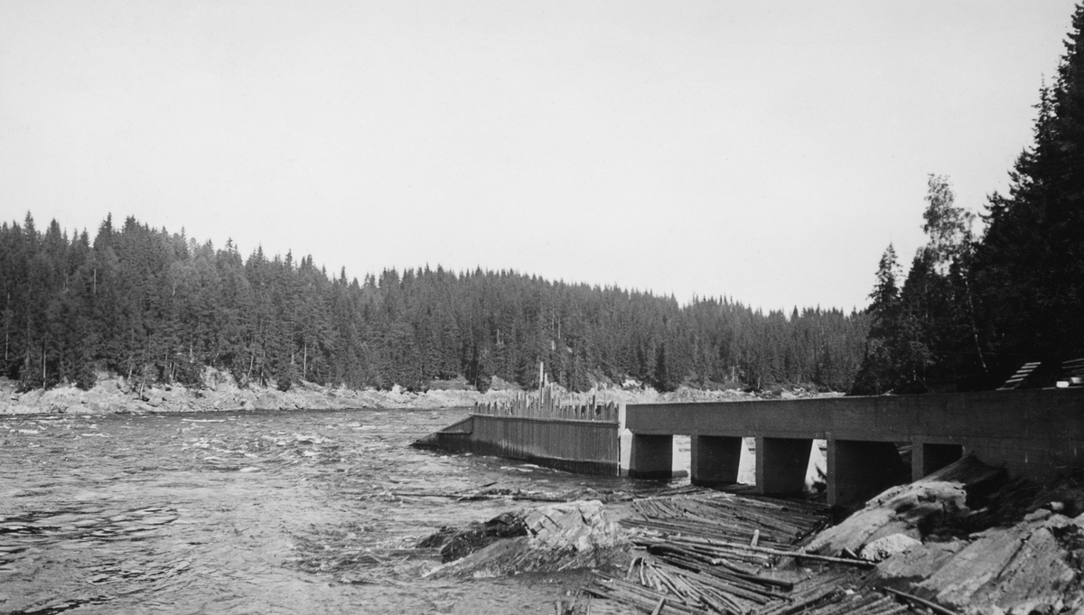 Utløpsdelen av den opprinnelige tømmerrenna (den som ble brukt i perioden 1916-26) ved Vamma kraftstasjon i Askim i Østfold.  Dette fotografiet er tatt i 1931.  Fire år tidligere, i 1927, var den avbildete renna erstattet av en tømmerkanal med litt større kapasitet.  Den avbildete konstruksjonen - ei slags bru eller akvedukt av betong som var ført et stykke ut i Glommas hovedløp - var imidlertid intakt.  Dette fotografiet viser hvordan Fredrikstad Tømmerdirektion i 1931 stengte de åpne hvelvingene under konstruksjonen, antakelig for å omskape brua til en skjerm som ledet tømmer som kom ovenfra mot den sentrale delen av elveløpet.  Her ser vi hvordan den ytre delen av brua på motstrøms side har fått en skjerm av tett, vertikalstilt plank.  Under de innerste brukarene er det imidlertid ikke bygd noen liknende skjerming, antakelig fordi denne delen av konstruksjonen lå i ly av et lite steinskjær som vi ser i forgrunnen på dette fotografiet.  Da bildet ble tatt lå det en del småtømmer på dette skjæret. 