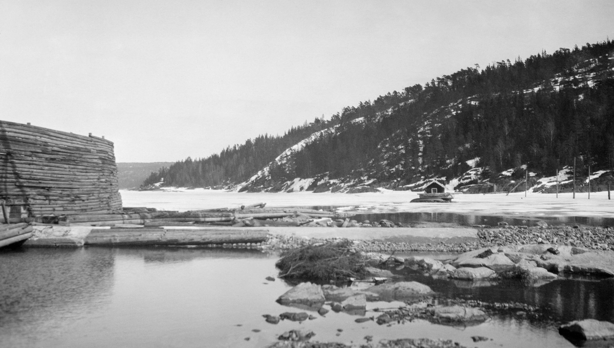 Fra Trøsken i Glommas vestre løp i Østfold. Fotografiet skal være tatt våren 1931, åpenbart så tidlig på våren at vårflommen ennå ikke hadde kommet. Til venstre ser vi en høy, buet skjerm, en type skådam som må har vært støttet av kraftige trebukker på landsida (baksida fra fotografens standpunkt). I det steinete løpet lå det en del tømmer, antakelig lensestokker. I forgrunnen var det en støpt terskel som dels var dekt med stein. I bakgrunnen til høyre lå det en flåte med ei hytte på, og framfor den lå en robåt hvelvet med kjølen oppovervendt.

Trøsken er et smalt sund mellom to vannspeil i den øvre delen av Glommas vestre løp i Østfold, Mingevannet (øverst) og Isnesfjorden. I dette sundet, som er bare om lag 30 meter bredt, ligger det steinur oppå en underliggende bergrygg. Det var bare i flomperioder at det var noenlunde enkelt å passere Trøsken med fløtingsvirke og båter. I en tilbakeskuende artikkel som sto i Sarpsborg Arbeiderblad 24. desember det året dette fotografiet ble tatt fortelles det at «… Det hendte ar folk gikk fra Skiptvedt til Sarpsborg og tilbake igjen, men det hørte til sjeldenhetene. Derimot gikk de som bodde ved Mingevannet og på denne side av Trøsken til byen. Og det de skulde selge og ha med sig tilbake igjen måtte de bære på ryggen eller med åk. Ved høivanne rodde man over Trøsken, ellers vadde man over. Sjelden var det heller ikke at man rodde ned Vestvannet og så bar varene videre til byen …».  Vinteren 1907-1908, mens man arbeidet med tømmertunnelen fra Svartdal («Toppen») ytterst i Isnesfjorden til Eidet ved Visterflo, ble det sprengt ut plass for et slusekammer ved Trøsken, men portene ble ikke montert. Etter hver kom man til at det ville være bedre å montere en klappedam i stedet. I 1930-åra ble problemet ved det trange og grunne sundet løst ved å bygge en ny dam ved Sølvstua, som stuvet opp såpass mye vann at passasjen ved Trøsken ble åpen for tømmer og små båter gjennom hele fløtingssesongen.