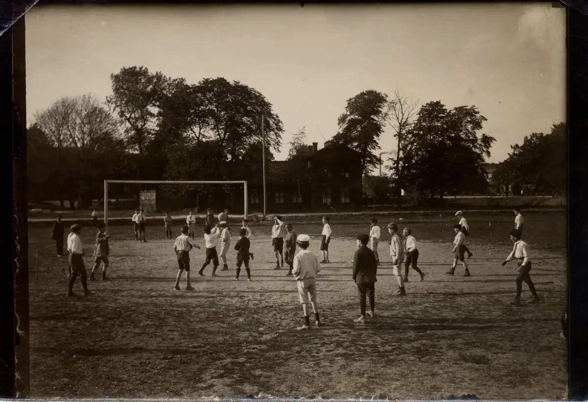 Ett trettiotal skolungdomar tränar fotboll. I bakgrunden fotbollsmål, väg, byggnad och grönska, 1910-talet.   Heden.