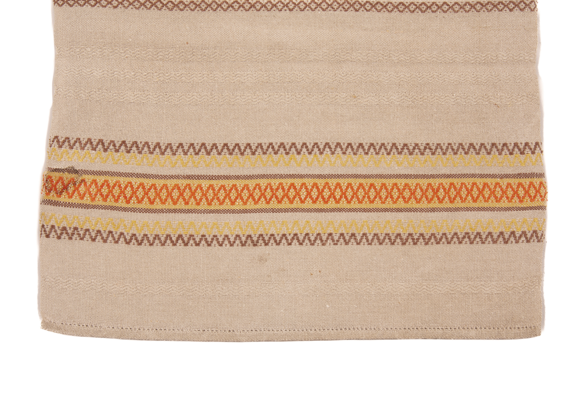 Beige løper med striper i stoffet, og border i endene. Faldet med hullsøm. Stripene er horisontale laget av et sikksakkmønster. Det er brune striper i en symmetrisk mønster langs hele duken, mellom de brune stripene er der tre parallelle horisontale lys beige striper. I hver ende er det en samling av striper i ulike farger som gul, rød og brun. 
