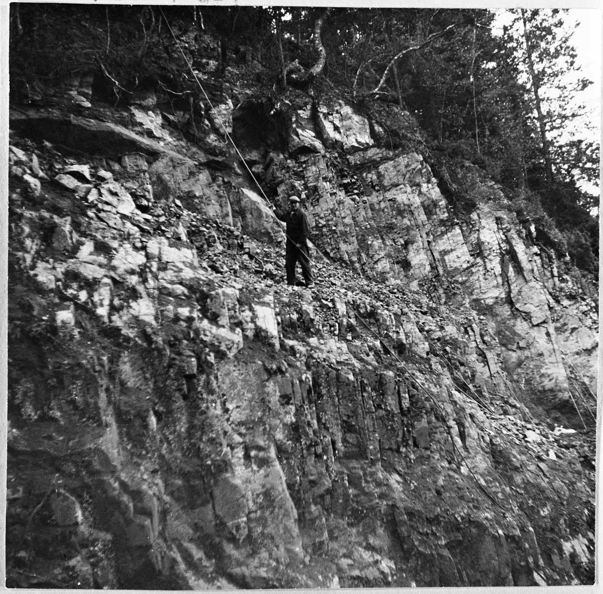 Bygging av skogsbilveg i Sullestadskogen i Totenvika ca. 1961-62. Her ser vi skytebas Osvald Halvorsen oppe i fjellskråningen der vegen skal gå. Han er sikret med et tau rundt livet. Man har fått sprengt ut ei lita hylle.