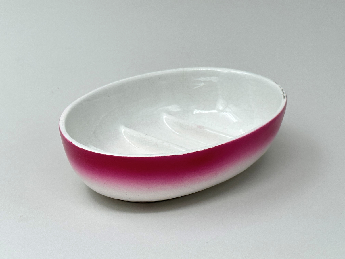 Oval tvålkopp i vitt flintgods med rosa inslag på utsidan. Botten tre gånger räfflad på både in- och undersida. 