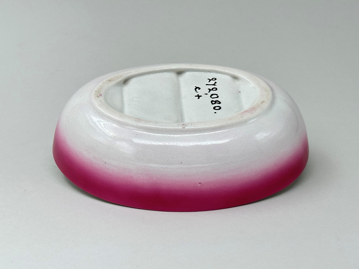 Oval tvålkopp i vitt flintgods med rosa inslag på utsidan. Botten tre gånger räfflad på både in- och undersida. 