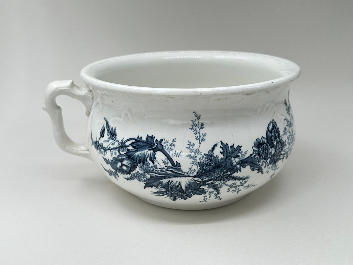 Rund potta i vitt porslin med blå dekor i form av blommor, blad och strån på fram- och baksida. Kanten något rundad. Dekorativt format handtag. 
