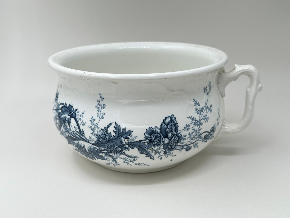 Rund potta i vitt porslin med blå dekor i form av blommor, blad och strån på fram- och baksida. Kanten något rundad. Dekorativt format handtag. 