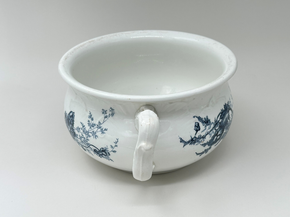 Rund potta i vitt porslin med blå dekor i form av blommor, blad och strån på fram- och baksida. Kanten något rundad. Dekorativt format handtag. 