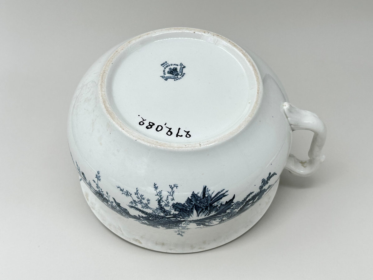 Rund potta i vitt porslin med blå dekor i form av blommor, blad och strån på fram- och baksida. Kanten något rundad. Dekorativt format handtag. 