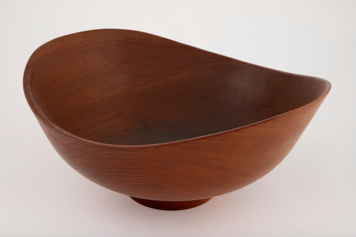 Skål i teak. Stor, dyp, oval form med liten, lav, sirkelrund fot. Skålen har skrånende sider som er høyest på kortsidene. Konsentriske årringer noe forskjøvet i forhold til skålens sentrum. På fotens underside er det tre gjenpluggede hull etter dreieskivens tapper.