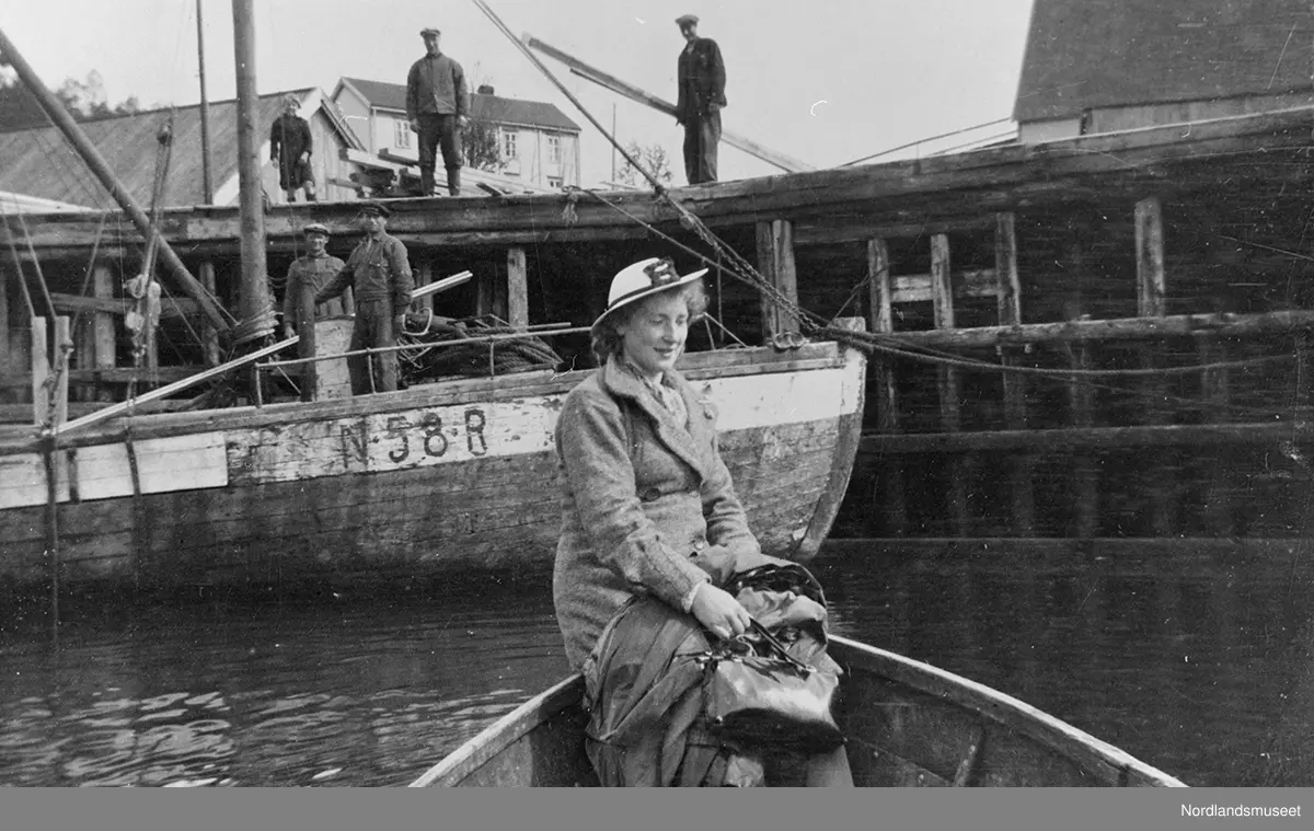 En kvinne, Anne Aarnes, sitter i baugen på en båt i forgrunnen. Bak henne ligger båten N-58-R "Binna" ved kai med to menn ombord. Oppå kaien bak står tre personer.