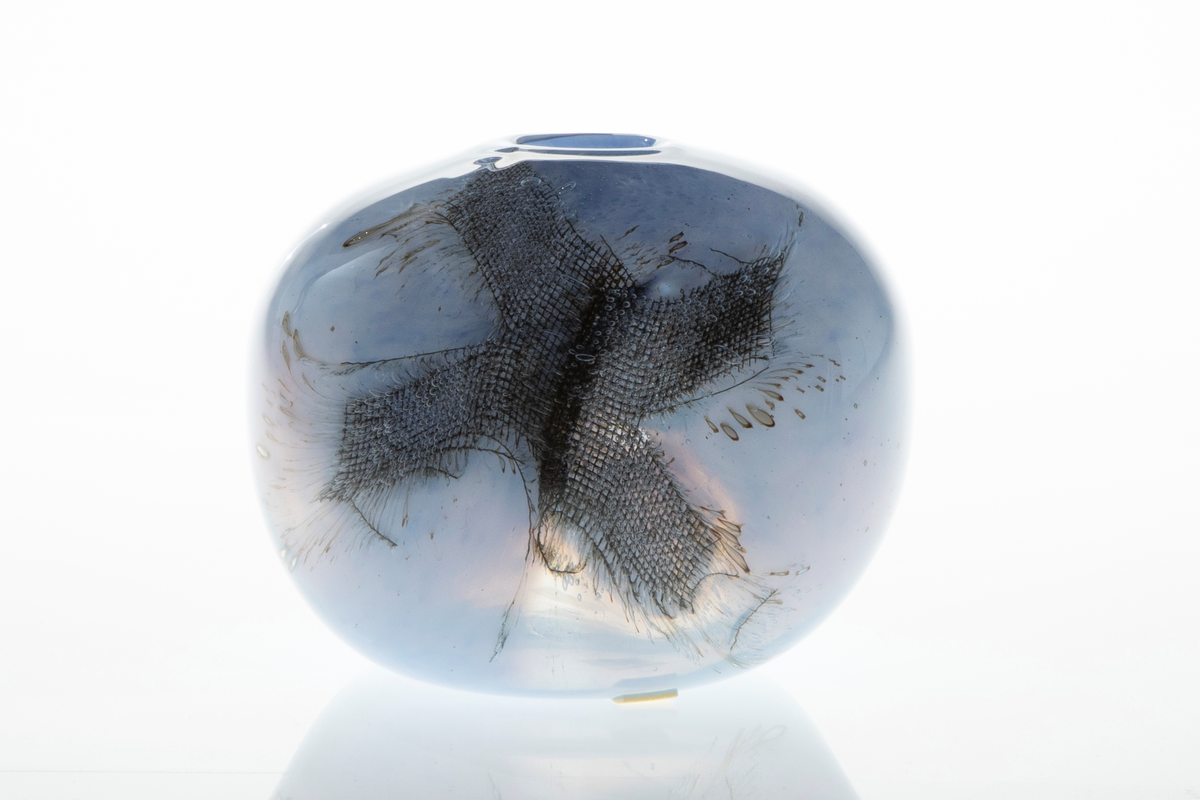 Kuleformet vase med liten rund munning i overfangsglass. Halvgjennomskinnelig melkehvitt glass med lyseblå fargepigmenter samt to innlagte metallnett. Sistnevnte danner et kryss, og ut ifra disse stråler lange, spisse luftbobler.