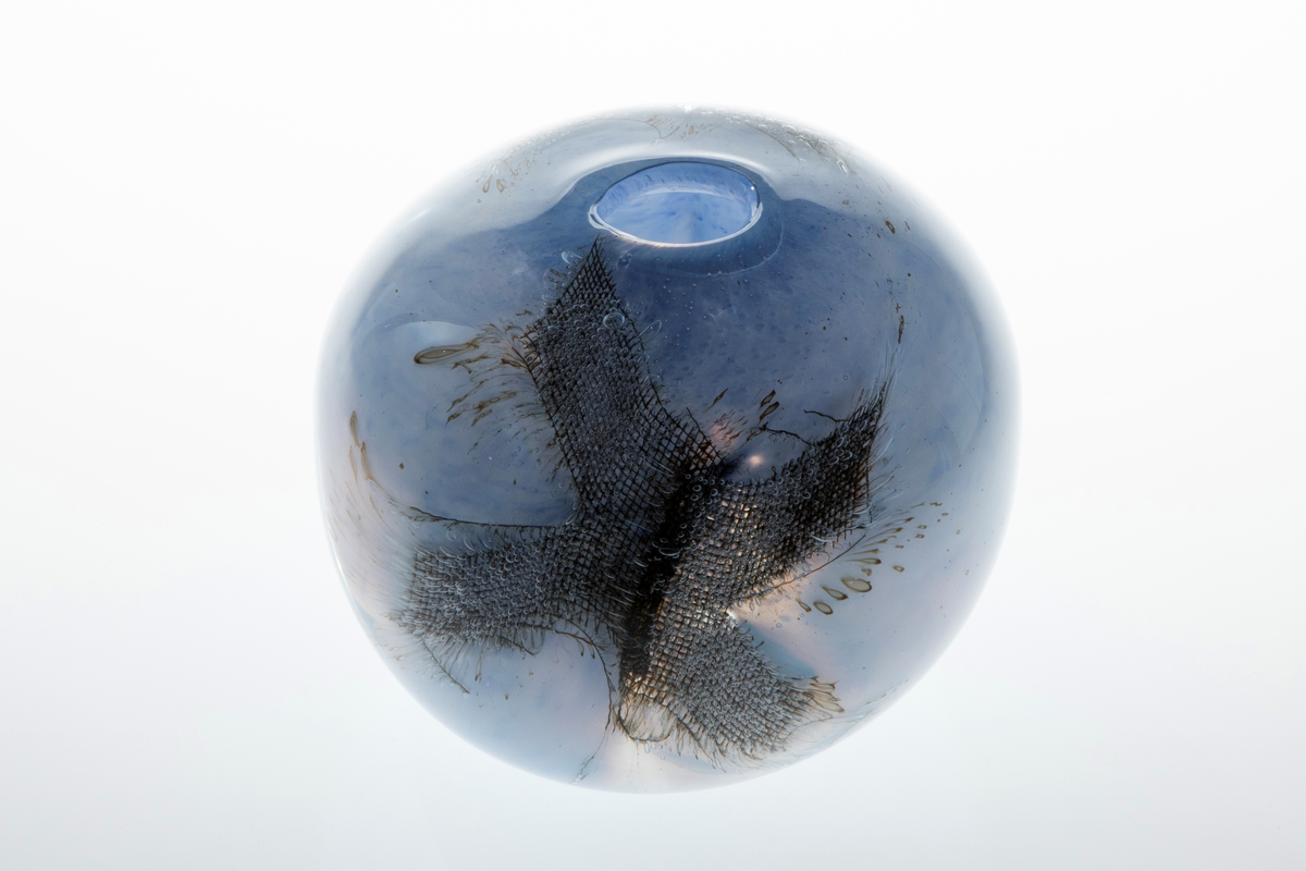 Kuleformet vase med liten rund munning i overfangsglass. Halvgjennomskinnelig melkehvitt glass med lyseblå fargepigmenter samt to innlagte metallnett. Sistnevnte danner et kryss, og ut ifra disse stråler lange, spisse luftbobler.