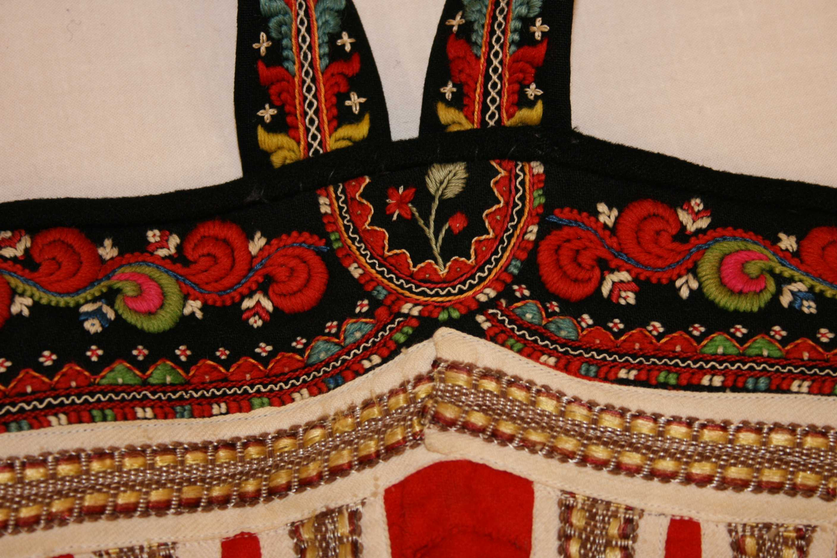 Svart vadmålsstakk med selar og broderi.