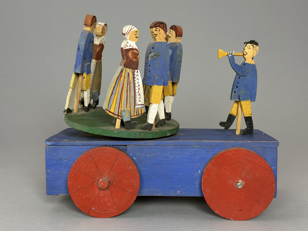 En blå kärra med röda hjul och grön dansbana med tre dansande par. Innuti ett "maskineri", som gör att banan snurras då kärran rullas.