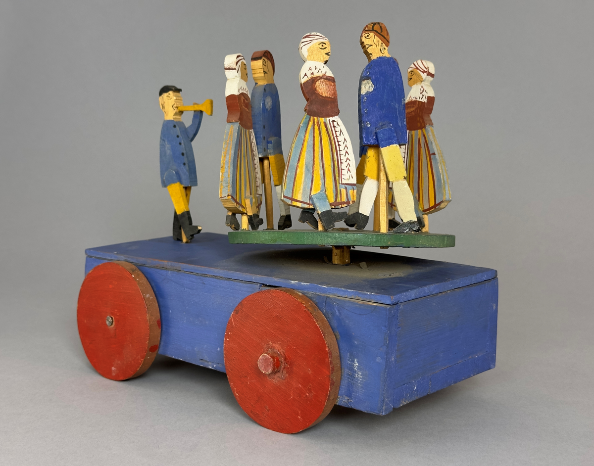 En blå kärra med röda hjul och grön dansbana med tre dansande par. Innuti ett "maskineri", som gör att banan snurras då kärran rullas.