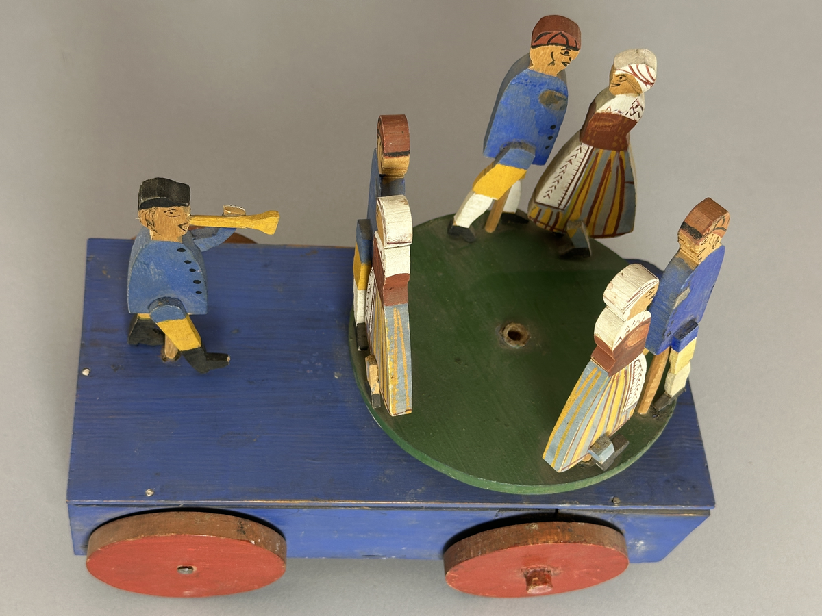 En blå kärra med röda hjul och grön dansbana med tre dansande par. Innuti ett "maskineri", som gör att banan snurras då kärran rullas.