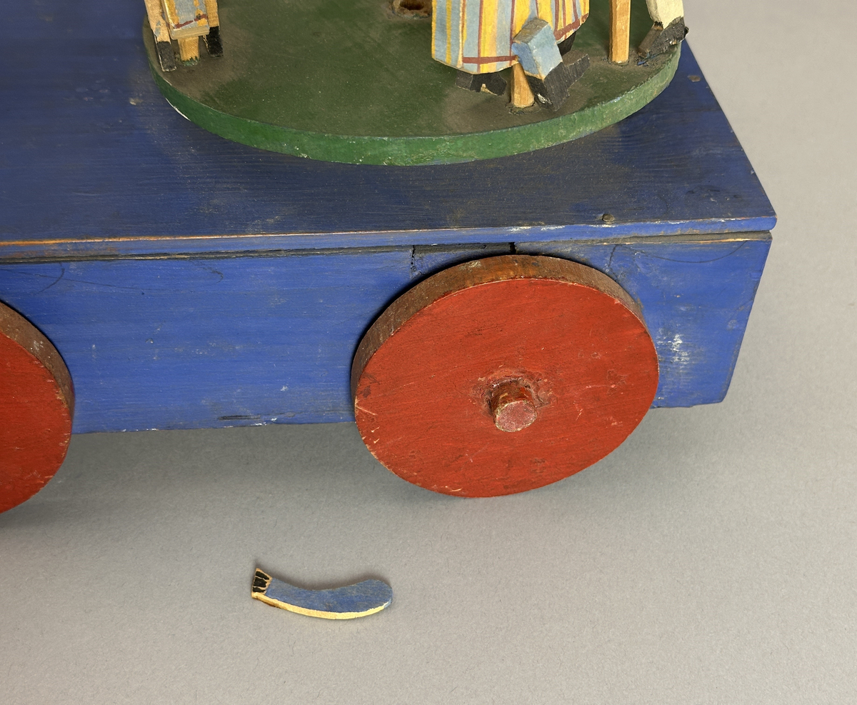 En blå kärra med röda hjul och grön dansbana med tre dansande par. Innuti ett "maskineri", som gör att banan snurras då kärran rullas.