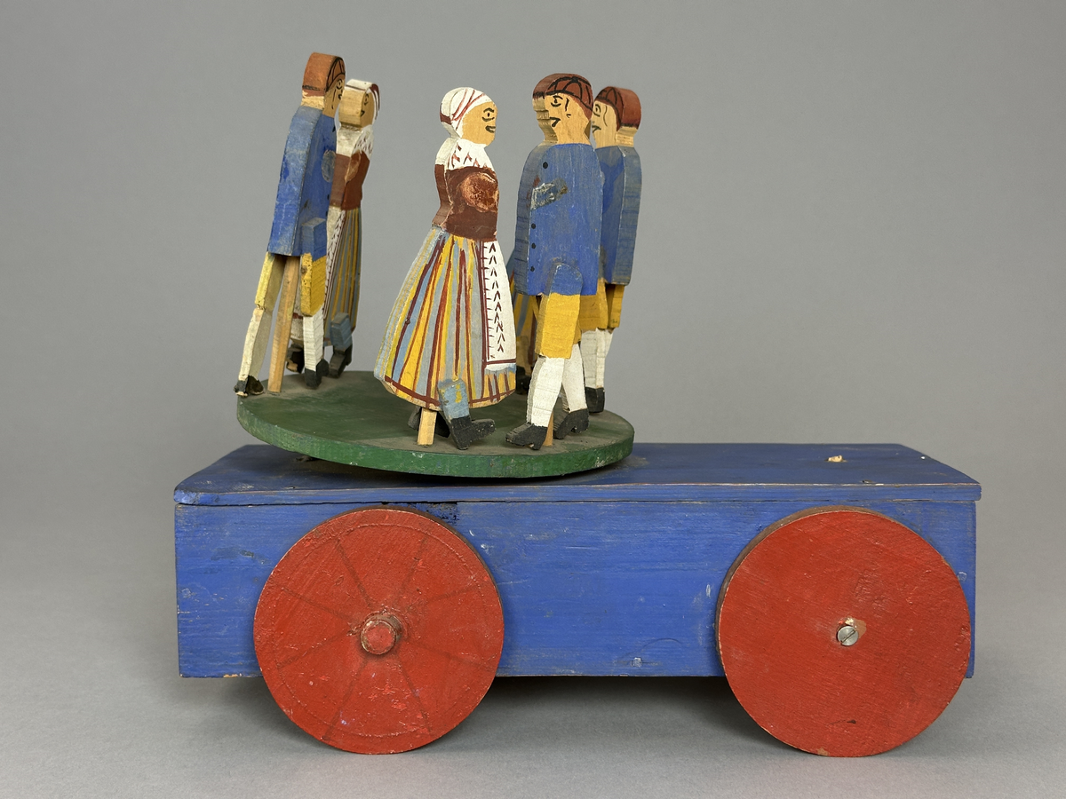 En blå kärra med röda hjul och grön dansbana med tre dansande par. Innuti ett "maskineri", som gör att banan snurras då kärran rullas.