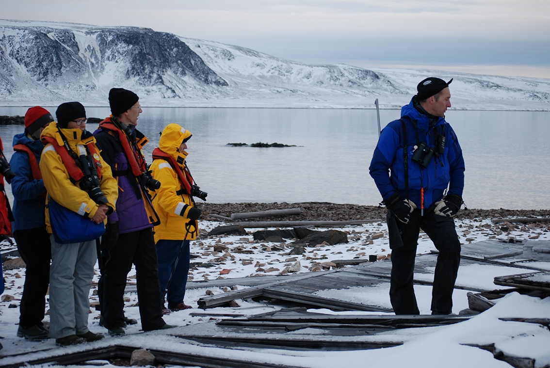 Turister och en guide med gevär över axeln vid en raserad byggnad.