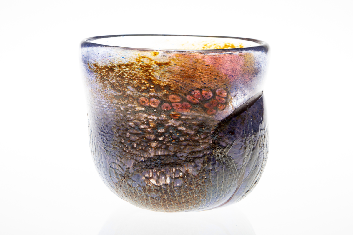 Tilnærmet sylindrisk bolle i halvgjennomskinnelig glass med iregulær form. På utsiden delvis ilagt nett. Fargesjatteringer i blå, lilla, og gulgrønn.