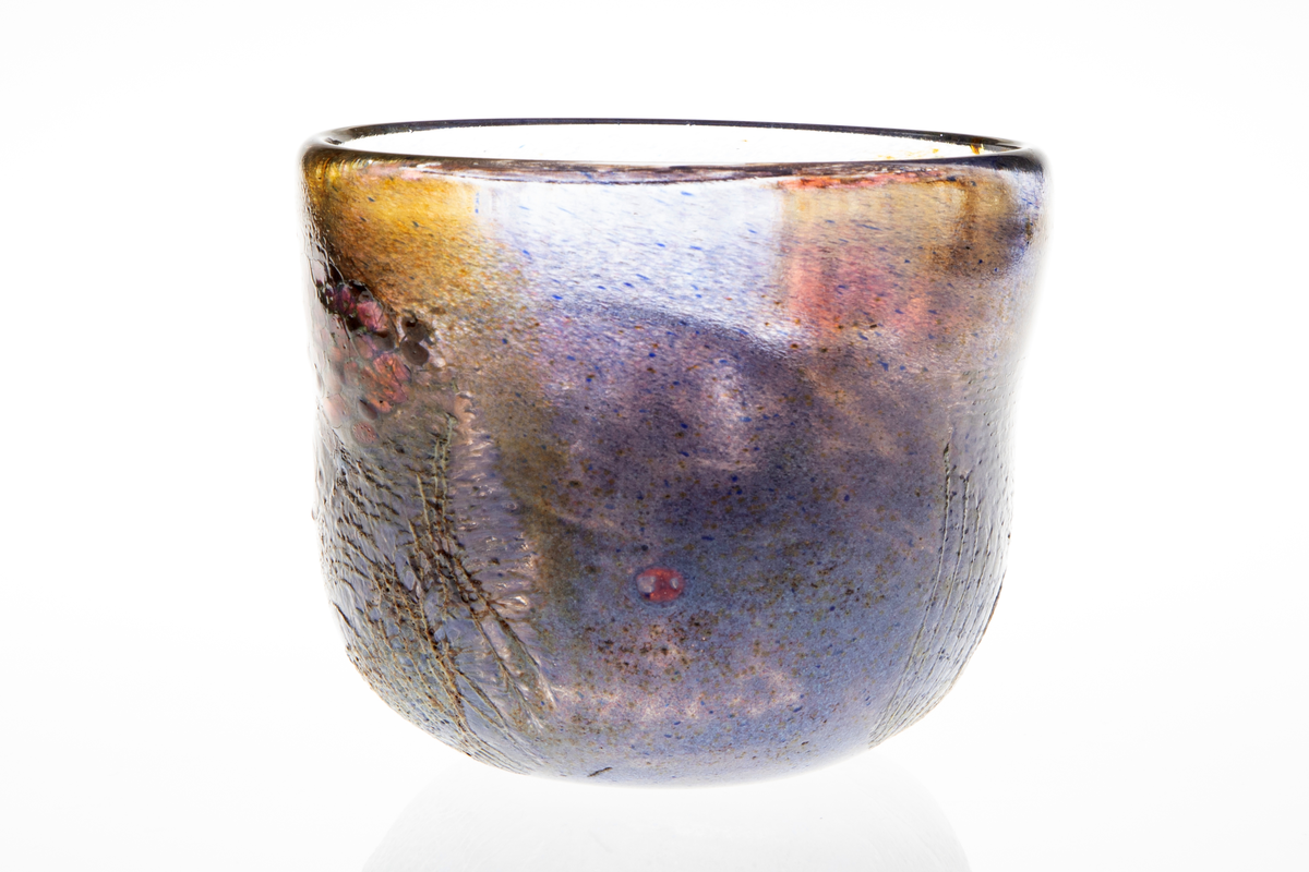 Tilnærmet sylindrisk bolle i halvgjennomskinnelig glass med iregulær form. På utsiden delvis ilagt nett. Fargesjatteringer i blå, lilla, og gulgrønn.