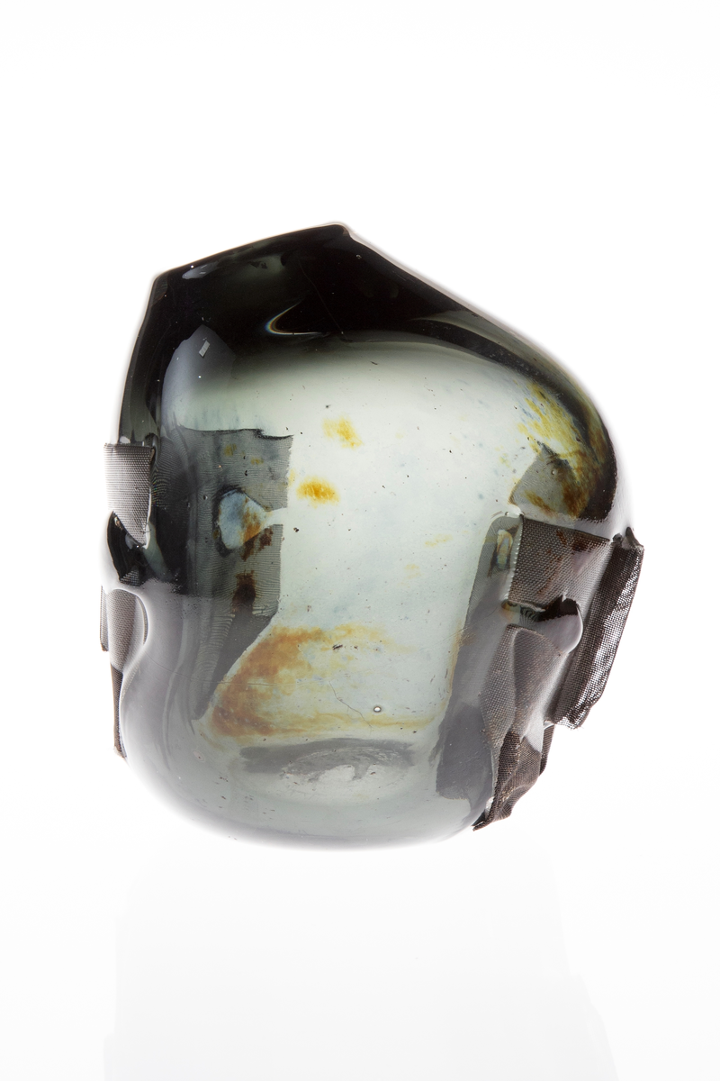 Vase med tilnærmet rund form. Rund åpning på en side i øvre del. Vasen har ilagte metallnett på to sider utformet som to ansikt