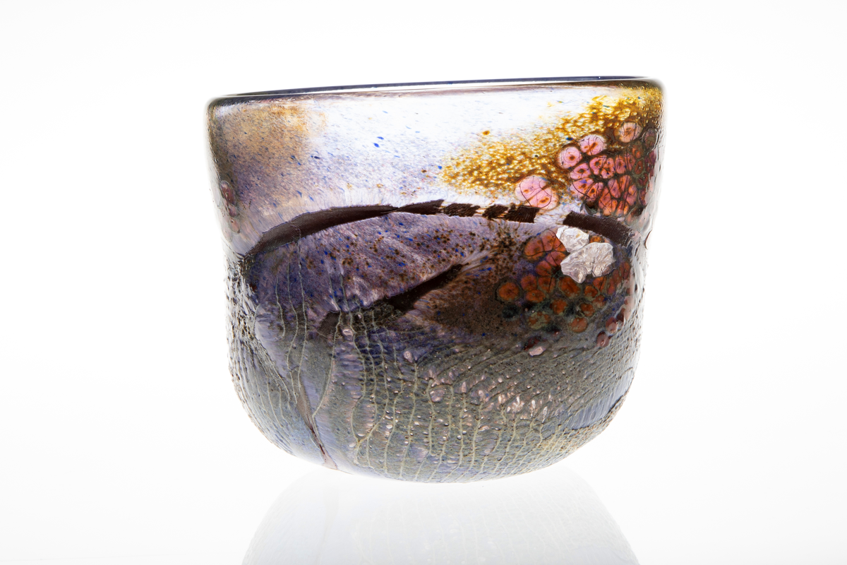 Tilnærmet sylindrisk bolle i halvgjennomskinnelig glass med iregulær form. På utsiden delvis ilagt nett. Fargesjatteringer i blå, lilla, og gulgrønn.