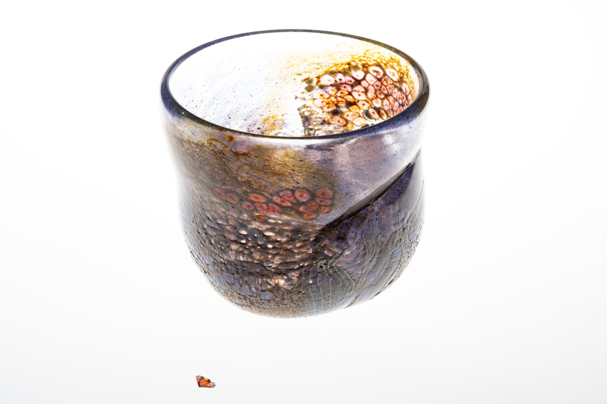 Tilnærmet sylindrisk bolle i halvgjennomskinnelig glass med iregulær form. På utsiden delvis ilagt nett. Fargesjatteringer i blå, lilla, og gulgrønn.