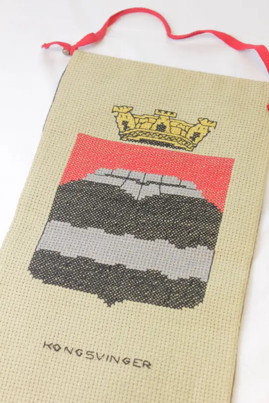 Pyntestreng med Kongsvinger kommunes byvåpen brodert i korssting på beige broderstoff, etter mønster fra en ukebladsserie "Våre vakre byåpen". Bakside av grått filtstoff. Opphenget er en messingstang med rødt bendelbånd. I bunnen henger to strikkede miniatyrvotter.