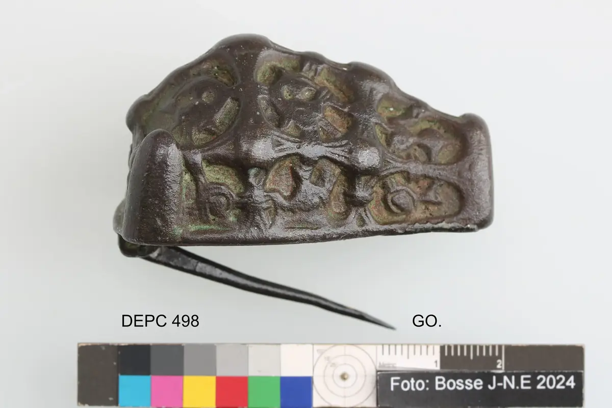 Spänne med ornamentik i kraftig relief, ovansidan indelad i fält med vulstförsedda lister, bottenplatta och nål bevarade, ganska nött
av brons, djurhuvudformat, längd 59, br. 36 h. 30 mm. 3 i längd - och 3 i tvåriktning gående med hög ornering prydda band, mellan dem kraftig ornering, (nålspetshylsan saknas). Förut märkt: 229.
