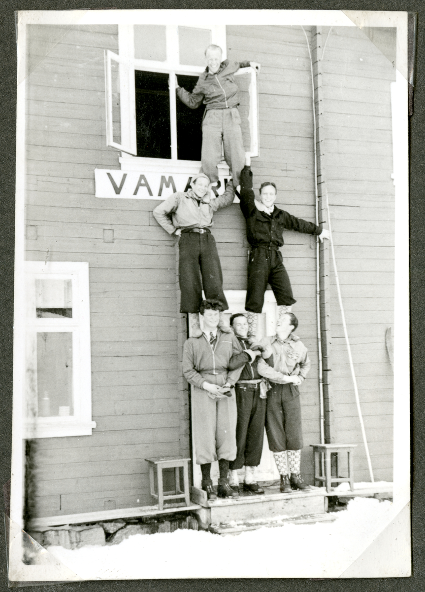 En vennegjeng skøyer på hyttetur der de har arrangert seg opp langs vegg i pyramideformasjon. Skilt på hytten: "Vamark"
