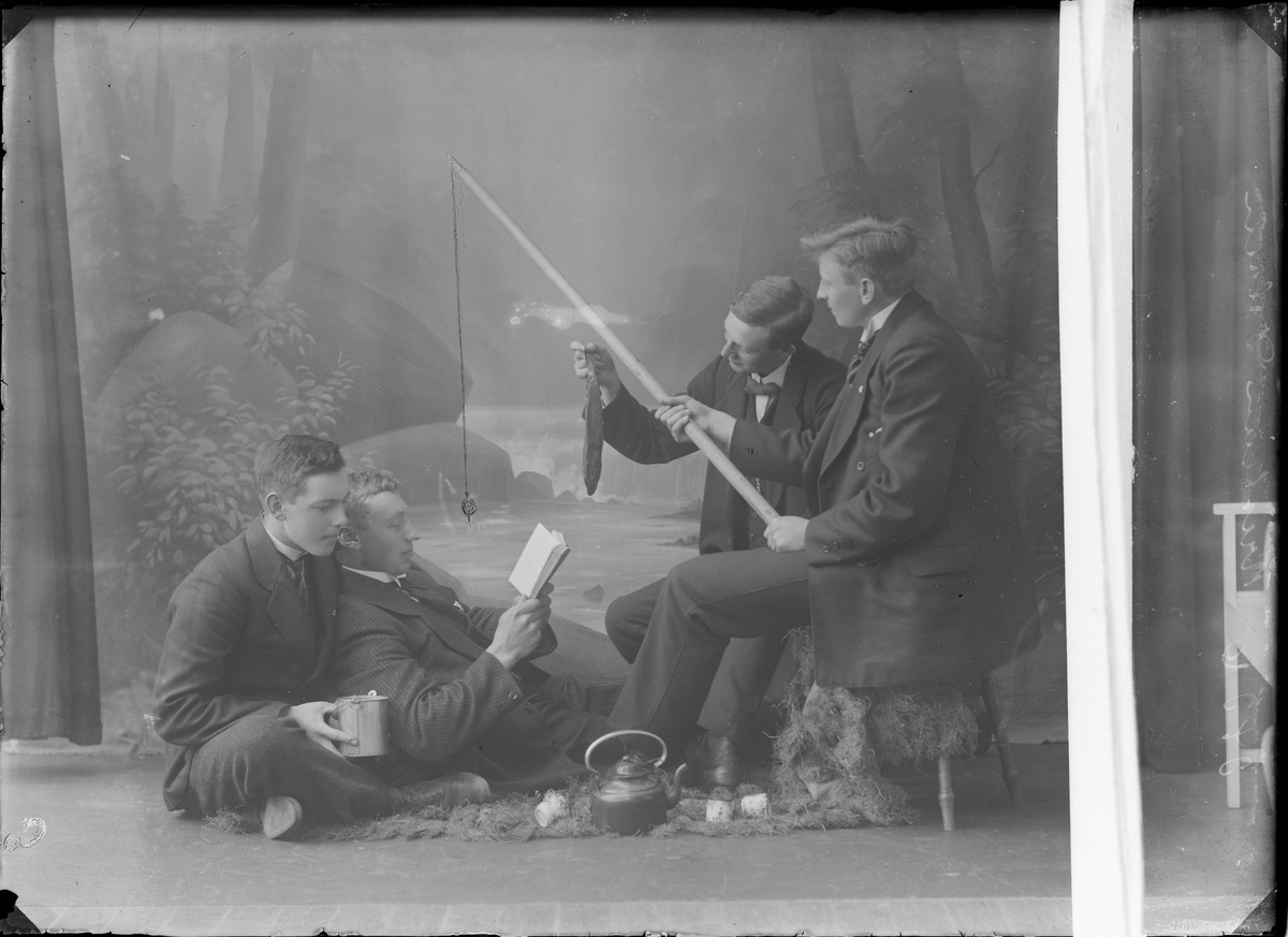 Gruppeportrett. Gruppe menn oppstilt i fotostudio med bålkjele, fisk, fiskestang og bok.
