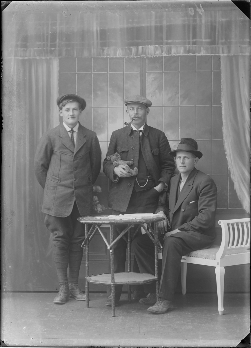 Gruppeportrett. Gruppe menn oppstilt i fotostudio. En av mennene har en kosebamse i armen og en tobakkspipe i munnen.